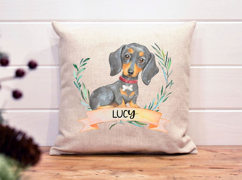 May include: A beige linen pillow with a watercolor illustration of a black and tan dachshund with a red collar and a bone charm. The dog is surrounded by green leaves and the name "LUCY" is written on a pink banner.