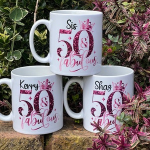 May include: Three white ceramic mugs with black handles are stacked. Each mug has a pink and glittery design that reads "Sis 50 & fabulous", "Kerry 50 & fabulous", and "Shaz 50 & fabulous". The mugs are decorated with a crown.