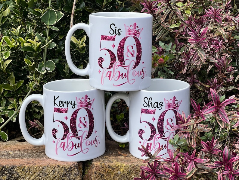 May include: Three white ceramic mugs with black handles are stacked. Each mug has a pink and glittery design that reads "Sis 50 & fabulous", "Kerry 50 & fabulous", and "Shaz 50 & fabulous". The mugs are decorated with a crown.
