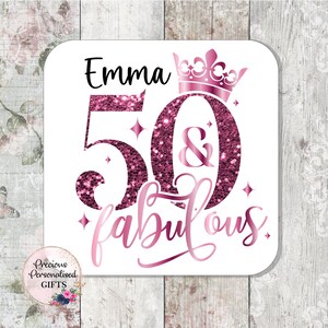 May include: A white square coaster with the name "Emma" in black, and the numbers "50" and the word "fabulous" in pink glitter. A pink crown sits above the number 0. The coaster has a floral design in the bottom left corner.
