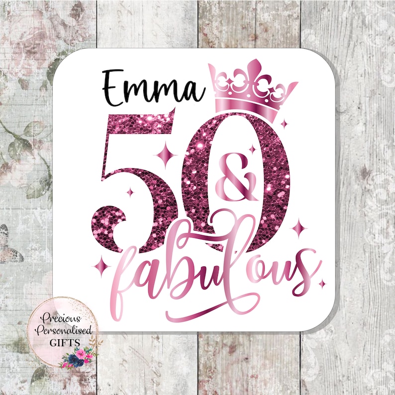 May include: A white square coaster with the name "Emma" in black, and the numbers "50" and the word "fabulous" in pink glitter. A pink crown sits above the number 0. The coaster has a floral design in the bottom left corner.
