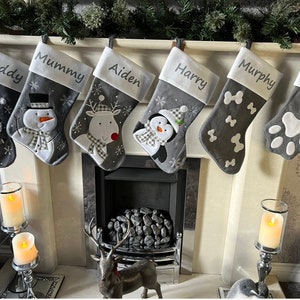 May include: Six grey Christmas stockings are displayed above a fireplace. Each stocking has a white cuff with a name embroidered on it: Daddy, Mummy, Aiden, Harry, Murphy, and Woody. The stockings are decorated with festive images including Santa, a snowman, and a penguin.