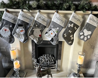 Puede incluir: Seis calcetines navideños grises cuelgan sobre una chimenea. Cada calcetín tiene un puño blanco con un nombre bordado: Daddy, Mummy, Aiden, Harry, Murphy y Woody. Los calcetines están decorados con imágenes festivas como Papá Noel, un muñeco de nieve y un pingüino.