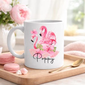 May include: White ceramic mug with a pink flamingo design and the name "Poppy" in cursive. The mug has a handle and is decorated with pink flowers and green leaves. The mug is on a wooden tray with a gold spoon.