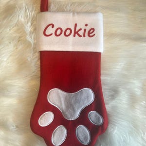 Pode incluir: Uma meia de Natal de feltro vermelho em forma de pata de animal com um punho branco. O punho branco apresenta o nome Cookie bordado em letras vermelhas. Uma al&ccedil;a de tecido vermelho est&aacute; presa no topo para pendurar a meia.