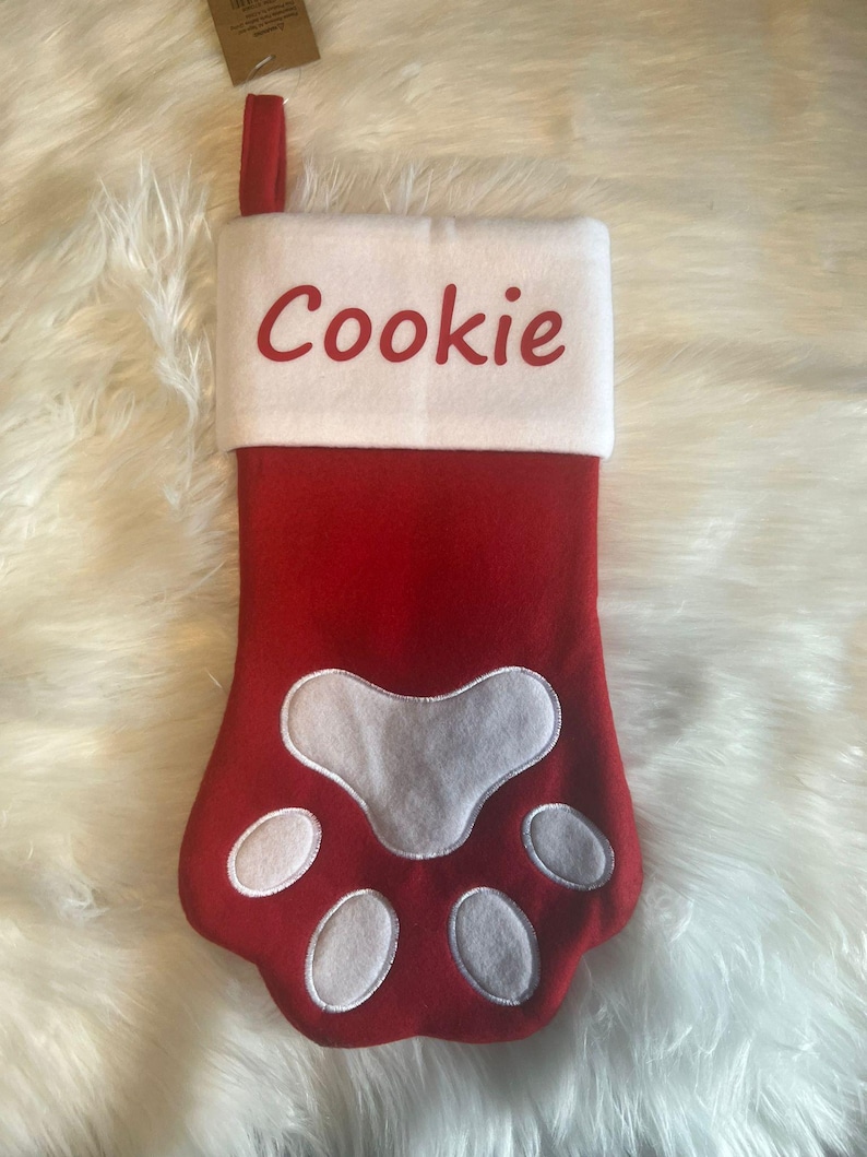 Pode incluir: Uma meia de Natal de feltro vermelho em forma de pata de animal com um punho branco. O punho branco apresenta o nome Cookie bordado em letras vermelhas. Uma al&ccedil;a de tecido vermelho est&aacute; presa no topo para pendurar a meia.