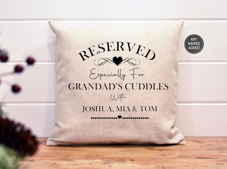 May include: A white pillow with black text that reads "RESERVED Especially For GRANDAD'S CUDDLES With JOSHUA, MIA & TOM". The text is surrounded by a decorative heart and scroll design.