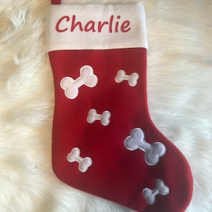 Pode incluir: Uma meia de Natal de feltro vermelho com um punho branco onde o nome Charlie est&aacute; impresso em letras vermelhas. O corpo da meia &eacute; decorado com v&aacute;rias formas de ossos de c&atilde;o bordadas em branco de tamanhos variados na parte frontal.
