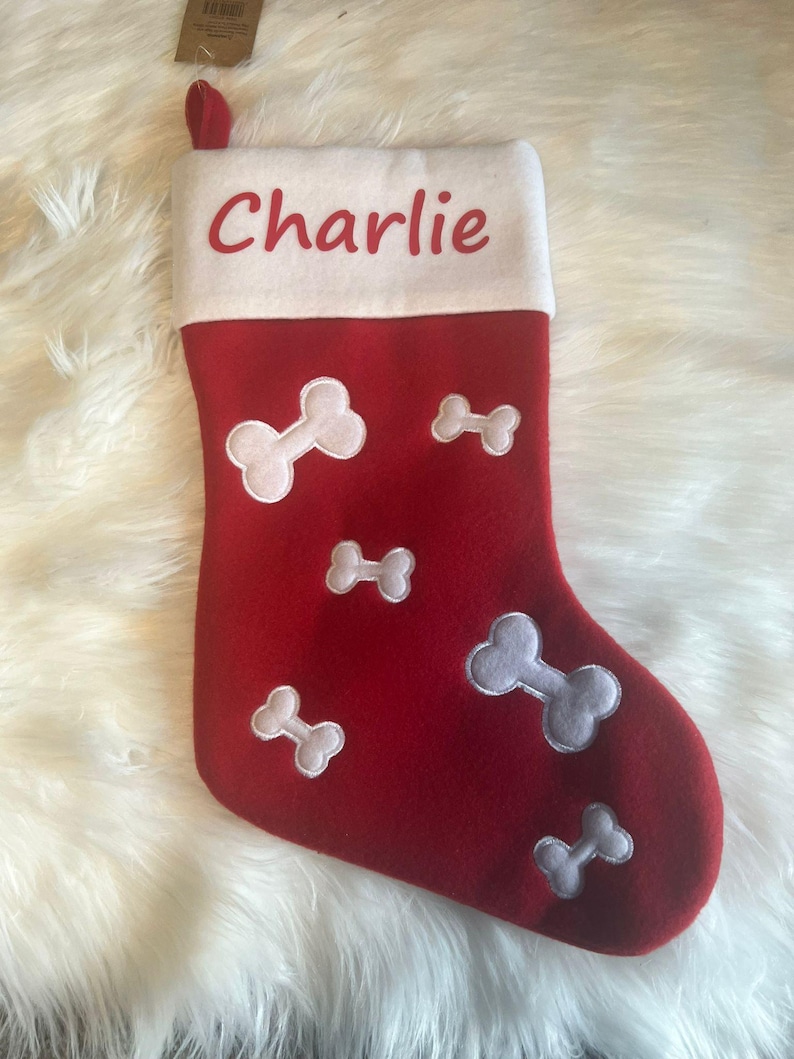 Pode incluir: Uma meia de Natal de feltro vermelho com um punho branco onde o nome Charlie est&aacute; impresso em letras vermelhas. O corpo da meia &eacute; decorado com v&aacute;rias formas de ossos de c&atilde;o bordadas em branco de tamanhos variados na parte frontal.