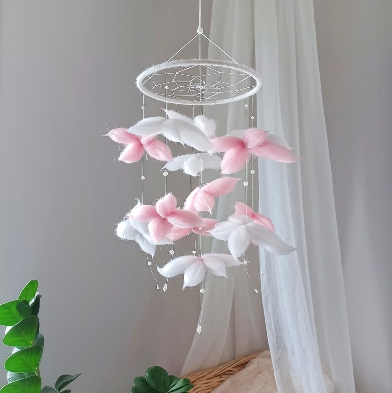 May include: A hanging mobile featuring a circular white frame with a webbed center. Suspended from the frame are multiple soft, fluffy flower-shaped elements in shades of white and pale pink, accented with small white beads on thin white strings.
