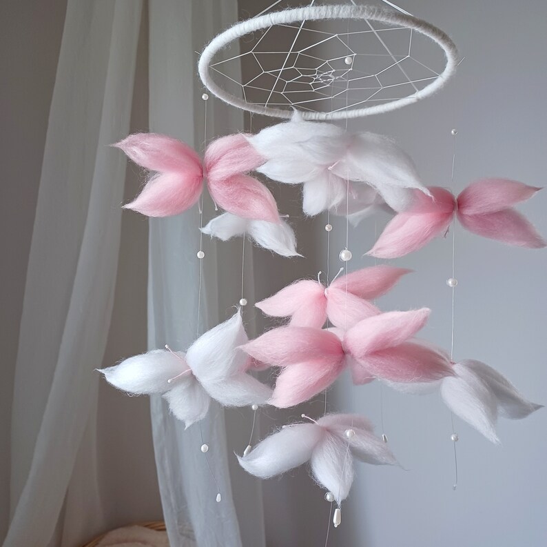 May include: A hanging mobile featuring a white circular frame with a webbed center. Suspended from the frame are multiple soft, fluffy butterfly-shaped elements in shades of white and light pink, accented with small white beads on thin strings.