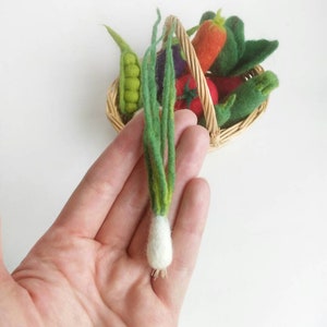 May include: A hand holding a felt green onion with a white bulb. The onion is part of a set of felt vegetables in a wicker basket.