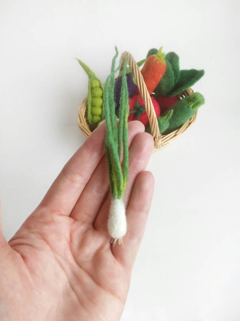 May include: A hand holding a felt green onion with a white bulb. The onion is part of a set of felt vegetables in a wicker basket.