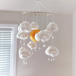 May include: A white felt cloud mobile with a yellow felt sun and crystal raindrops hanging from the clouds. The mobile is suspended from a gold metal ring.