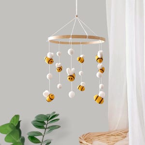 May include: A hanging mobile featuring a circular wooden frame with white strings suspended from it. Attached to the strings are several felted wool bees with yellow and black stripes, along with small white felted balls hanging at various heights.