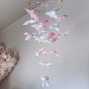 May include: A hanging mobile featuring a circular gold frame with a white webbed center. Numerous soft, fluffy white and light pink cloud-like shapes are suspended from the frame by thin strings, interspersed with small, round, white beads.