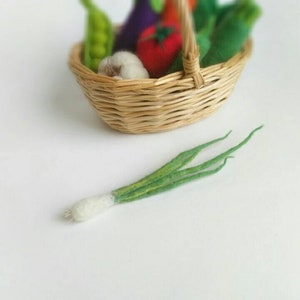 May include: A wicker basket filled with felt vegetables, including a green eggplant, a red tomato, a white garlic, and green beans. A green felt onion is lying on a white surface next to the basket.