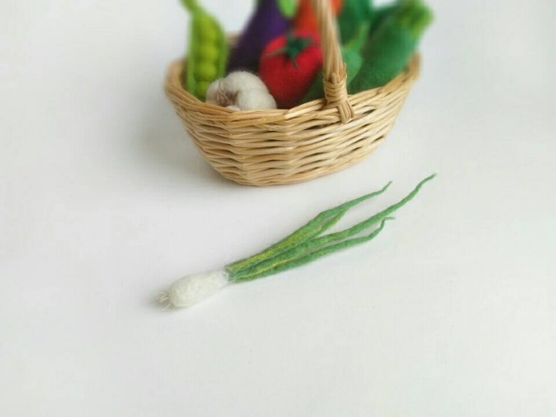 May include: A wicker basket filled with felt vegetables, including a green eggplant, a red tomato, a white garlic, and green beans. A green felt onion is lying on a white surface next to the basket.