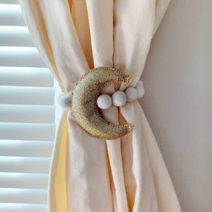 May include: A gold crescent moon curtain tieback with white pom poms. The tieback is attached to a white curtain.