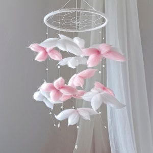 May include: A hanging mobile featuring a circular white frame with a webbed center. Suspended from the frame are multiple white and soft pink fiber flower-shaped elements interspersed with small white beads on thin, clear hanging strings.