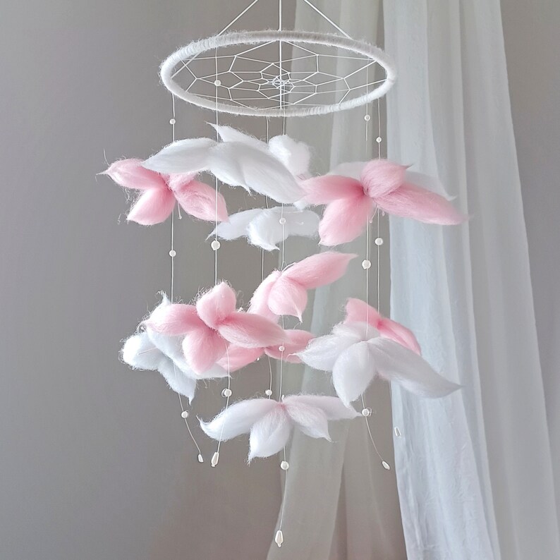 May include: A hanging mobile featuring a circular white frame with a webbed center. Suspended from the frame are multiple white and soft pink fiber flower-shaped elements interspersed with small white beads on thin, clear hanging strings.