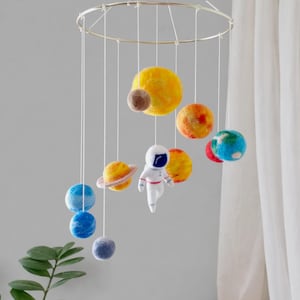 May include: A handmade solar system mobile featuring felt planets in shades of yellow, orange, blue, and brown, suspended from a gold ring. Includes a white astronaut figure. A decorative nursery or playroom accent.