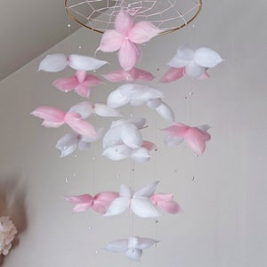 May include: A hanging mobile featuring a gold-toned circular frame with a webbed center. Suspended from the frame are multiple soft, fluffy flower-shaped elements in shades of white and light pink, interspersed with small white beads on thin strings.