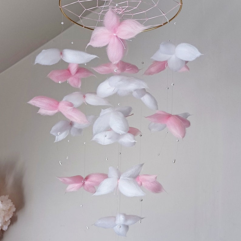 May include: A hanging mobile featuring a gold-toned circular frame with a webbed center. Suspended from the frame are multiple soft, fluffy flower-shaped elements in shades of white and light pink, interspersed with small white beads on thin strings.