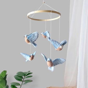 May include: A hanging mobile featuring four needle-felted birds suspended from a circular wooden hoop by thin white strings. Each bird has light blue wings and a tan-coloured chest, depicted in various flight positions with wings spread wide.