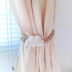 Könnte beinhalten: Ein weißer, wolkenförmiger Vorhanghalter mit einer Schnur aus beigefarbenen Filzkugeln. Der Vorhanghalter ist um einen cremefarbenen Vorhang gewickelt und bildet einen dekorativen Akzent. Der Vorhang befindet sich neben einem Fenster mit weißen Jalousien.