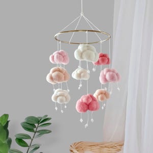May include: A nursery mobile with felt clouds in pink, cream, and peach hues. The clouds are suspended from a gold-toned ring with clear crystal beads. This decorative mobile is designed for a baby's room or a child's bedroom.
