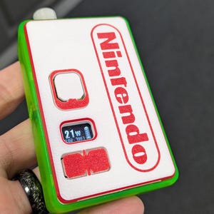May include: A green and white handheld device with a red Nintendo logo and a small screen displaying "21W".