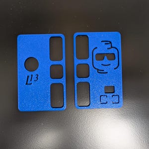 May include: Two blue rectangular phone case covers. One has the text "L13" and a circular cutout. The other features a cartoon face with sunglasses and several rectangular cutouts. The cases are made of a textured material.