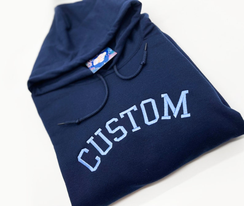May include: A navy blue hooded sweatshirt with the word "CUSTOM" embroidered in light blue on the front.