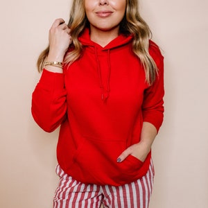 May include: A red hooded sweatshirt with a kangaroo pocket. The sweatshirt is worn by a person with long blonde hair and is paired with red and white striped pants.