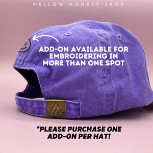May include: Purple baseball cap with a small embroidered smiley face. The text "ADD-ON AVAILABLE FOR EMBROIDERING IN MORE THAN ONE SPOT" is printed on the cap. The text "*PLEASE PURCHASE ONE ADD-ON PER HAT!" is printed below the cap.