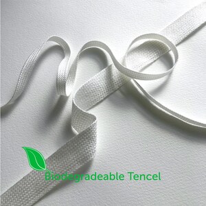 Może przedstawiać: Zbliżenie białej, tkanej wstążki z zielonym tekstem "Biodegradable Tencel". Wstążka jest zakrzywiona i zapętlona, podkreślając jej fakturę i kształt na białym tle. Materiał wydaje się miękki i elastyczny.