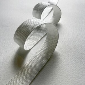 May include: A length of white woven ribbon, looped and curved against a white background. The ribbon has a textured, basket-weave pattern. The lighting creates shadows and highlights, emphasizing the ribbon's form and texture. The ribbon appears to be made of a durable material.