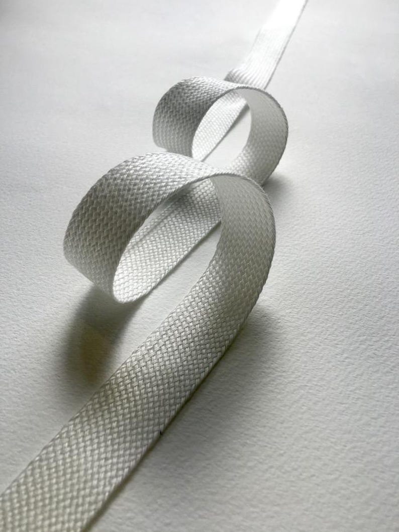 May include: A length of white woven ribbon, looped and curved against a white background. The ribbon has a textured, basket-weave pattern. The lighting creates shadows and highlights, emphasizing the ribbon's form and texture. The ribbon appears to be made of a durable material.