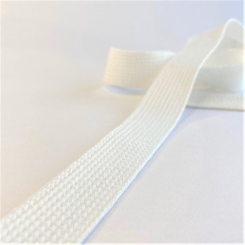 May include: Close-up of a length of white woven ribbon. The ribbon has a textured surface and is slightly curved, with a portion of the ribbon rolled up in the background. The ribbon is likely used for sewing, crafting, or decorative purposes.