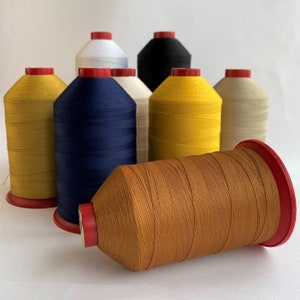 Puede incluir: Surtido de carretes de hilo en varios colores, incluyendo amarillo, azul, blanco, marrón y negro. Los carretes están dispuestos en semicírculo sobre una superficie blanca.