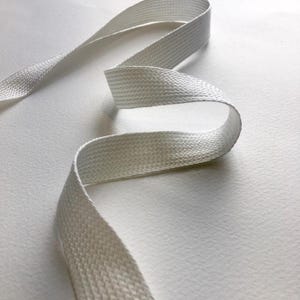 May include: A length of white, woven ribbon, gracefully curved across a white surface. The ribbon's texture is visible, with a subtle pattern. The overall composition is simple and clean, highlighting the ribbon's form and texture.