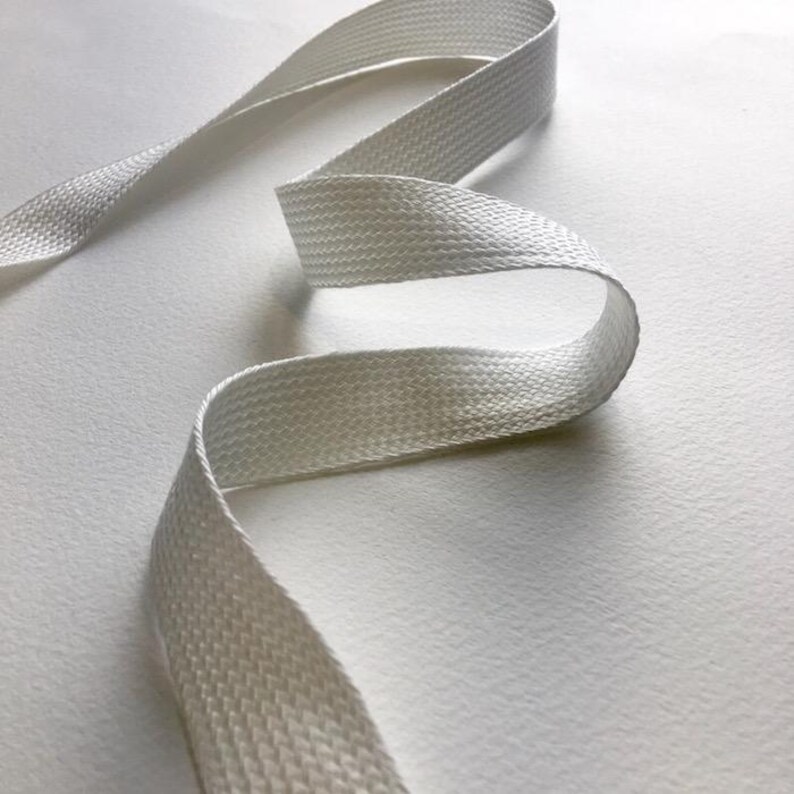 May include: A length of white, woven ribbon, gracefully curved across a white surface. The ribbon's texture is visible, with a subtle pattern. The overall composition is simple and clean, highlighting the ribbon's form and texture.