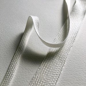 May include: Close-up of a white, woven ribbon with a textured surface. The ribbon is folded and curved, creating a dynamic visual effect. The weave pattern is visible, and the ribbon appears to be made of a durable material. The background is a soft, light-colored surface.