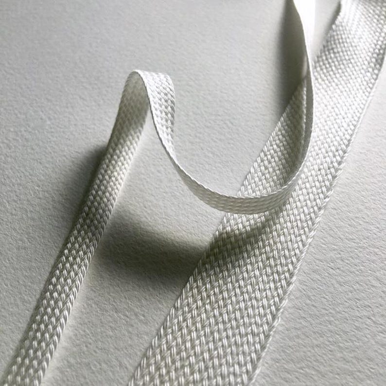 May include: Close-up of a white, woven ribbon with a textured surface. The ribbon is folded and curved, creating a dynamic visual effect. The weave pattern is visible, and the ribbon appears to be made of a durable material. The background is a soft, light-colored surface.
