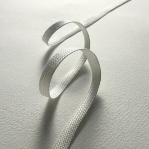 Könnte beinhalten: Ein weißer, flacher Schnürsenkel mit gewebtem Muster. Der Schnürsenkel ist in einer Spirale vor einem weißen Hintergrund aufgerollt, wodurch ein Gefühl von Bewegung und Textur entsteht. Der Schnürsenkel ist etwa 1,3 cm breit.