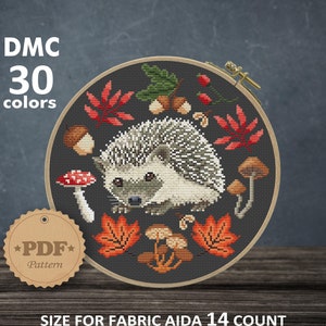 May include: Cross stitch pattern featuring a hedgehog surrounded by autumn leaves, mushrooms, and acorns. The pattern is for Aida 14 count fabric and measures 8 inches by 8 inches, or 20.32 cm by 20.32 cm. The pattern requires 30 DMC colours.