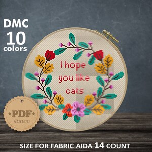 Puede incluir: Patrón de punto de cruz con una corona de flores y el texto "Espero que te gusten los gatos". El patrón está diseñado para tela Aida de 14 hilos y mide 6,93 x 6,43 pulgadas o 17,60 x 16,33 cm.