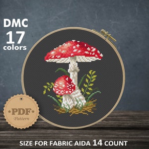 May include: Cross stitch pattern of a red and white mushroom with white spots, a smaller red and white mushroom, and green leaves on a dark gray background. The pattern is in a hoop and the text "DMC 17 colors" and "*PDF* Pattern" is visible. The size of the pattern is 10.16 x 13.24 cm or 4.00 x 5.21 inches and uses 56 x 73 stitches.