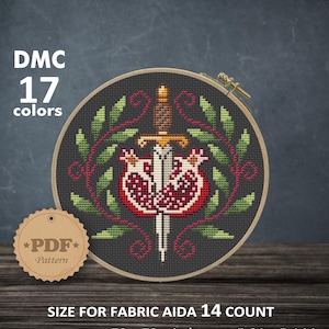 May include: A cross-stitch pattern in a circular wooden hoop featuring a central dagger piercing a pomegranate, surrounded by green leaves and red vines. Text includes DMC 17 colors, PDF Pattern, and size for fabric Aida 14 count 5.21 x 5.14 inches.